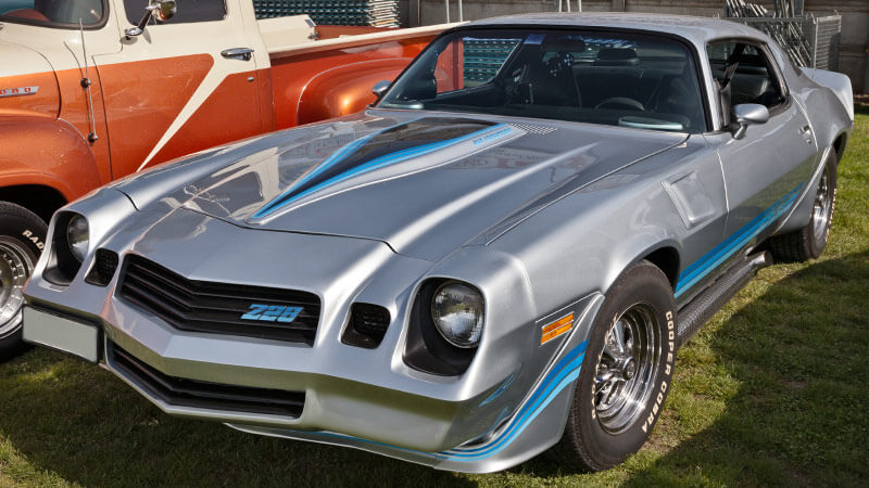 A silver 1970-1981 Chevy Camaro with blue and black decals on the hood and sides parked in a grass field