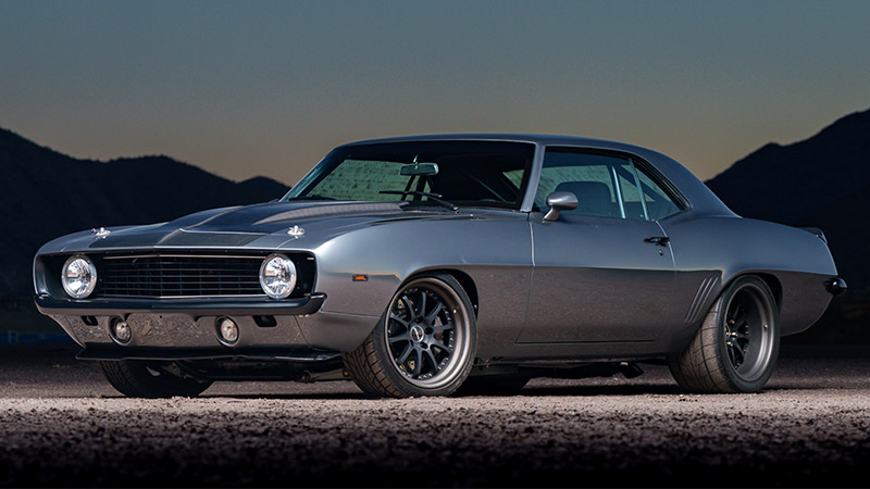 A wide angle shot of a gray classic Chevy Camaro with black racing stripes parked on in a parking lot in front of a dark background