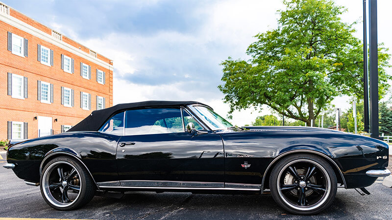 A wide angle shot of the side of a black 1967 Chevy Camaro convertible with black aftermarket wheels parked in a parking lot with a brick building in the background