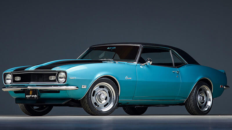 A wide angle shot of the side of a blue 1968 Chevy Camaro with black racing stripes parked in a showroom