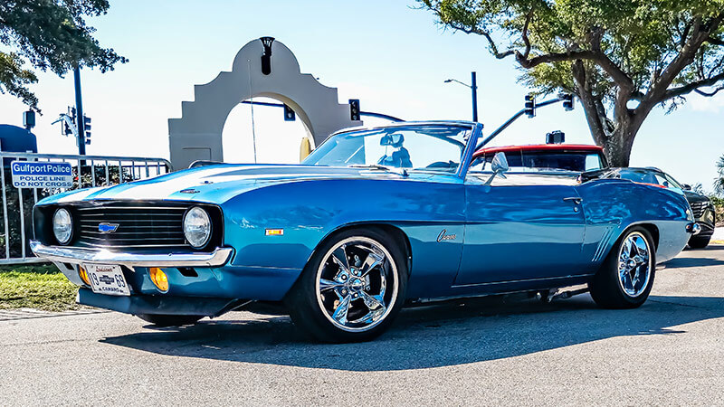 A wide angle shot of the side of a blue 1969 Chevy Camaro COPO convertible parked in front of a gray stone arch with trees in the background