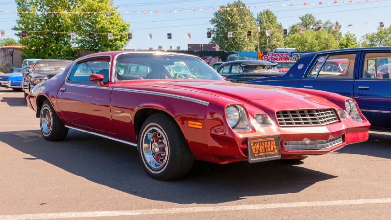 A wide angle shot of a red 1979 Chevy Camaro with silver wheels parked in a parking lot at a car show