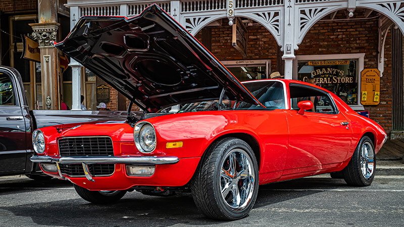 A wide angle shot of a red classic Chevy Camaro with the hood open at a car show parked next to a pickup truck with a brick building in the background