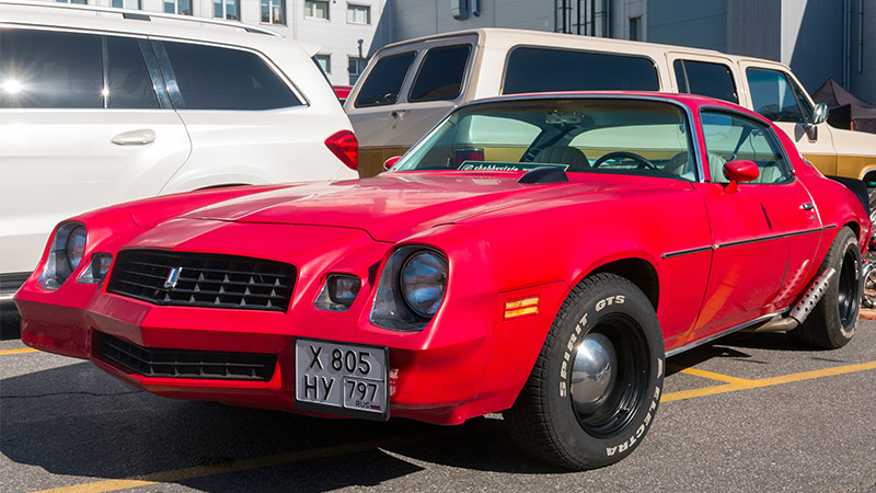 A wide angle shot of a red 1977 Chevy Camaro with black wheels parked in a parking lot at a car show