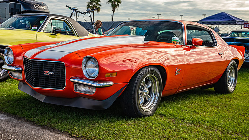 A wide angle shot of an orange 1973 Chevy Camaro Z/28 with silver wheels parked in a grass field at a car show