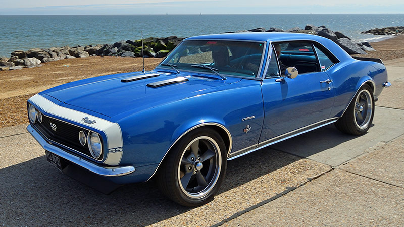 A blue classic Chevy Camaro with silver accents parked on a sun-bleached road with a rocky beach in the background