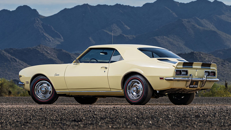 A tan classic Chevy Camaro with black racing stripes parked on a gravel lot with a large mountain in the background