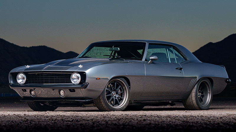 A silver classic Chevy Camaro with black racing stripes parked in a gravel lot with a large mountain in the background