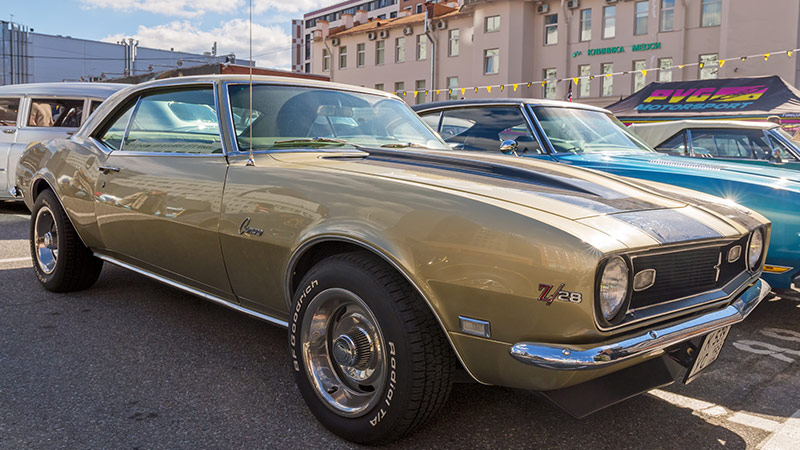 A beige classic Chevy Camaro parked in a parking lot at a car show with other classic Chevys