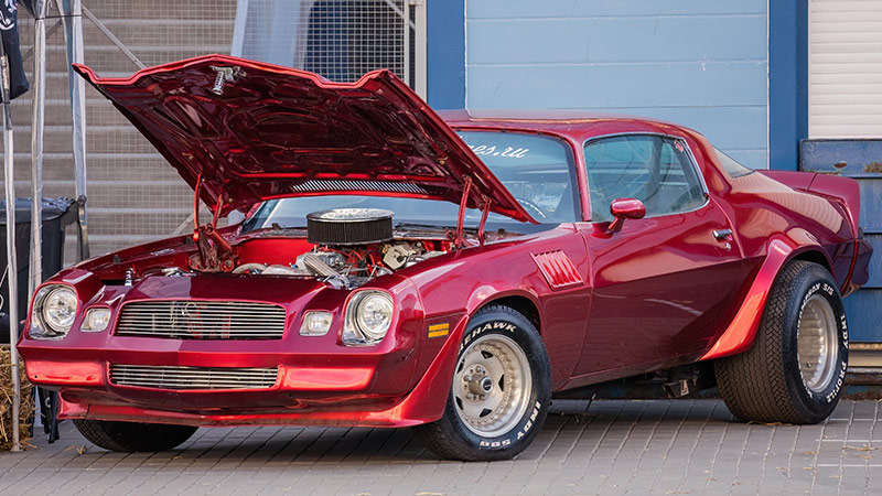 A red classic Chevy Camaro with large aftermarket gold wheels with its hood popped open while parked