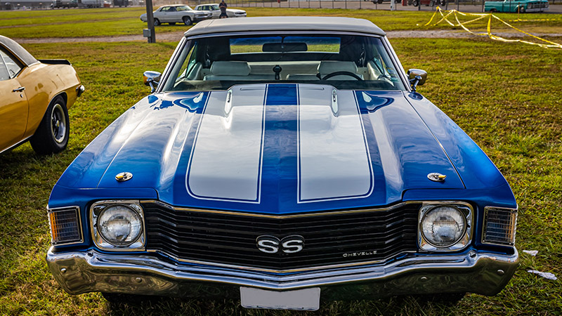 A close up of the front end of a blue 1972 Chevy Chevelle with white racing stripes parked in a field of grass